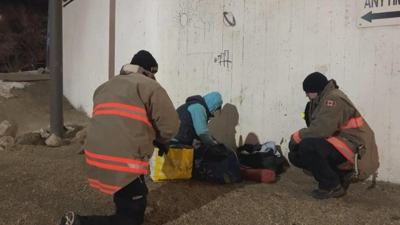A woman in a light blue jacket sitting next to two fire fighters knelt on the ground.