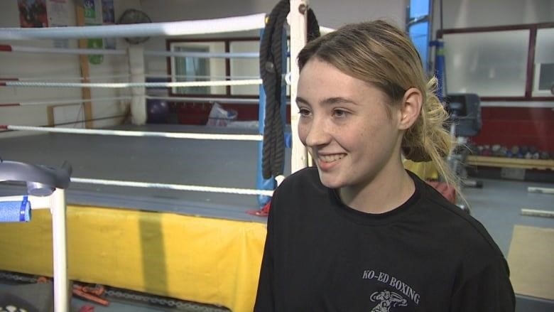 Young woman with blonde hair tied back in ponytail stands in a boxing gym wearing black t-shirt.