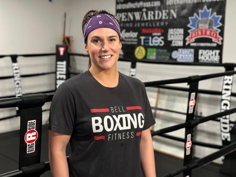 A woman wearing a boxing Tshirt stands next to a boxing ring 
