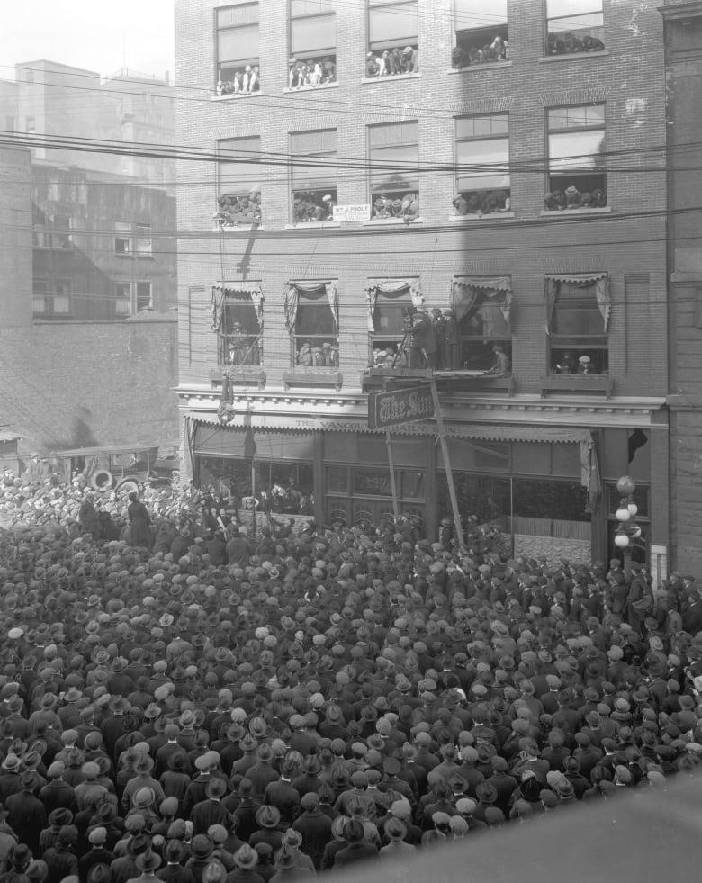A crowd of people forms around the Vancouver Sun Building, Houdini is suspended upsidedown middair in a straightjacket. 