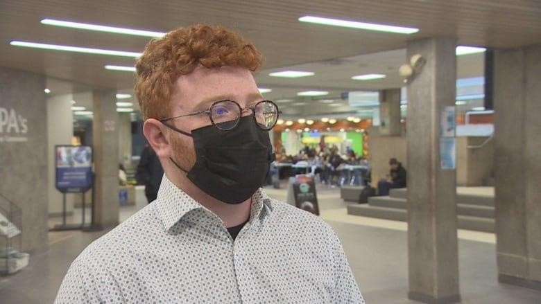 A man with curly red hair and glasses stands in a hallway with a mask on.