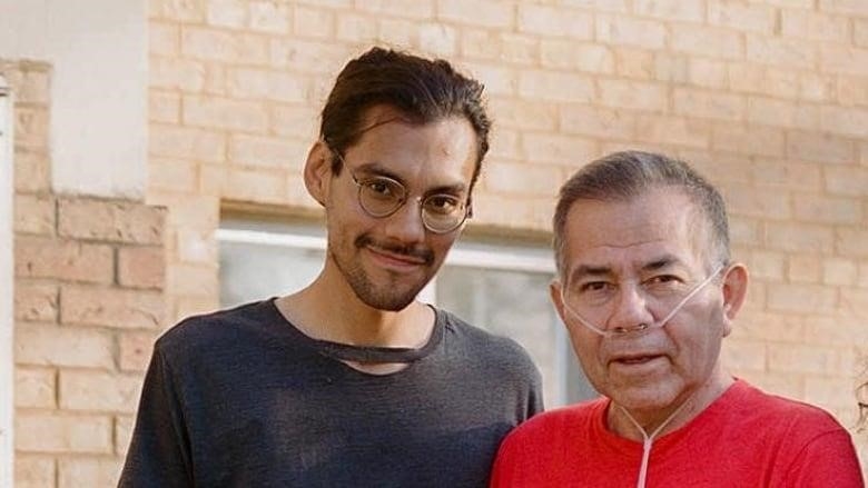 A younger smiling man stands next to a man wearing an oxygen tube. 