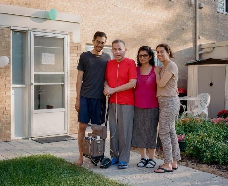 Four smiling people stand closely together in an embrace. One of the men wears an oxygen tube and stands next to a trolley carrying an oxygen machine. 