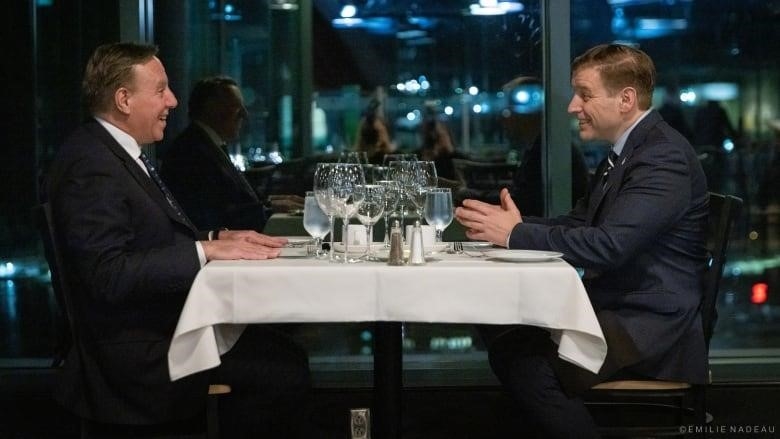 Two men in suits sit across a dinner table from each other, both smiling, with plates and wine glasses in between them.