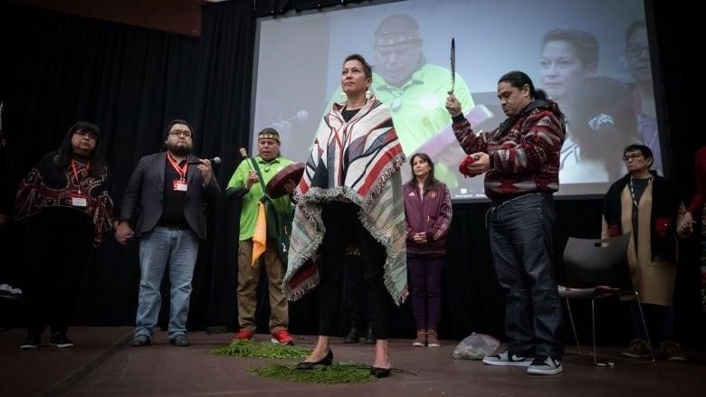 An Indigenous woman stands in the middle of a circle of First Nations leaders, wrapped in a blanket. An Indigenous man holds up a feather and a pouch.
