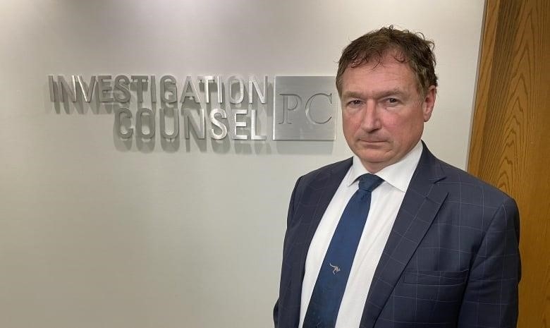 A man stands in front of a wall with the name of his law firm written on it.