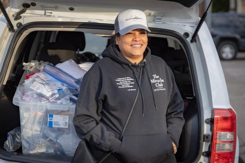 A Black woman smiles in front of an open car trunk with medical supplies visible.