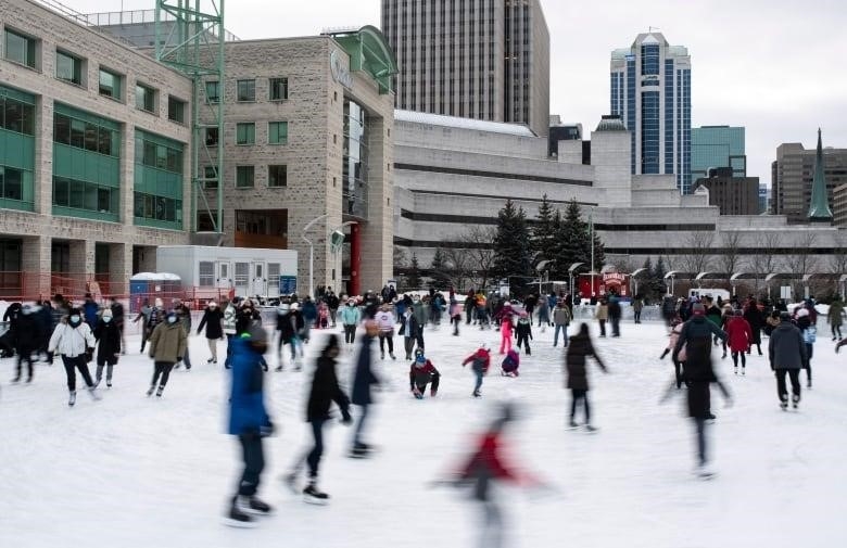 An outdoor skating rink next to a city hall and courthouse. Some skaters have a motion blur.