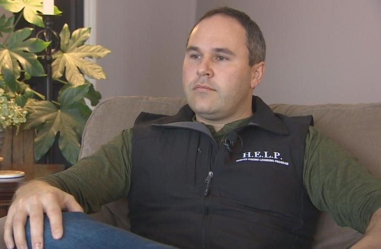 A man rests his arm on his knee. He is wearing a black vest with a H.E.L.P. logo on the side. 