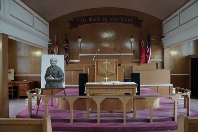 A view of the inside of a small church, with a portrait of a Black woman on the left, red carpeting and wooden architecture. 