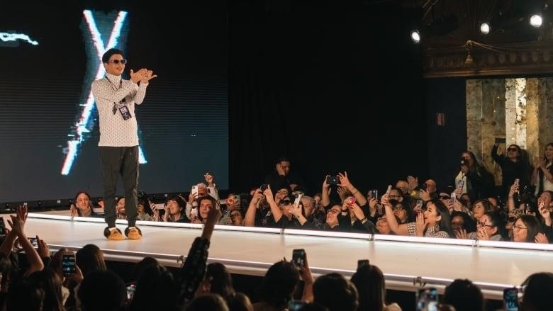 Person stands on fashion runway with hands palm up in front of audience.