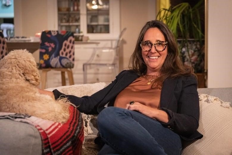 A woman with long brown hair and glasses sits on a couch with a dog and a big smile. 