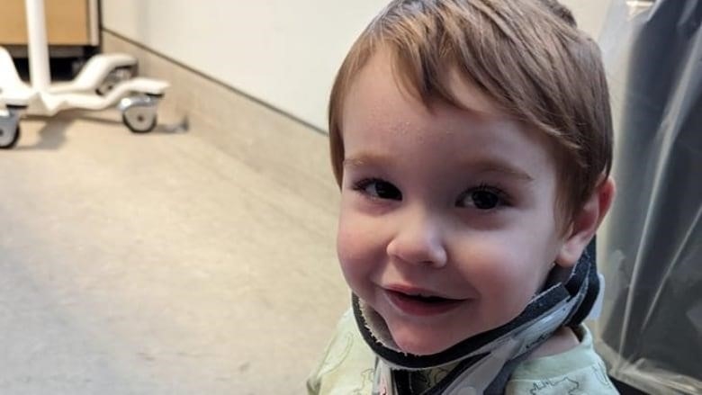 A toddler boy sits on the floor and smiles at the camera. He is wearing a neck brace. 