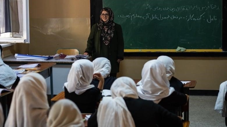 Female students in white headscarves are seen from behind sitting at desks, as a teacher standing by a blackboard smiles at them from the front of the class.