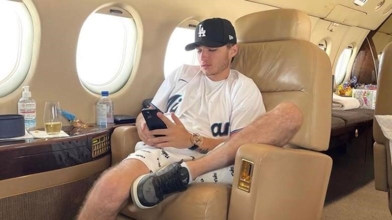 A young man sits in the seat of a private jet looking at his phone. 