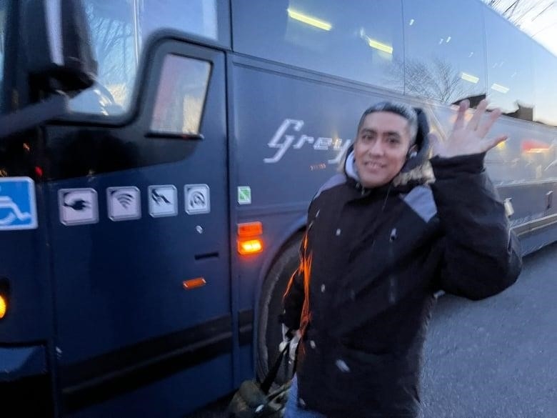 A man waves at the camera, a Greyhound bus in the background. 