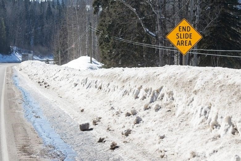 A yellow diamond-shaped highway sign that says "END SLIDE AREA" is pictured on the side of a snowy highway. A forest is visible in the background.