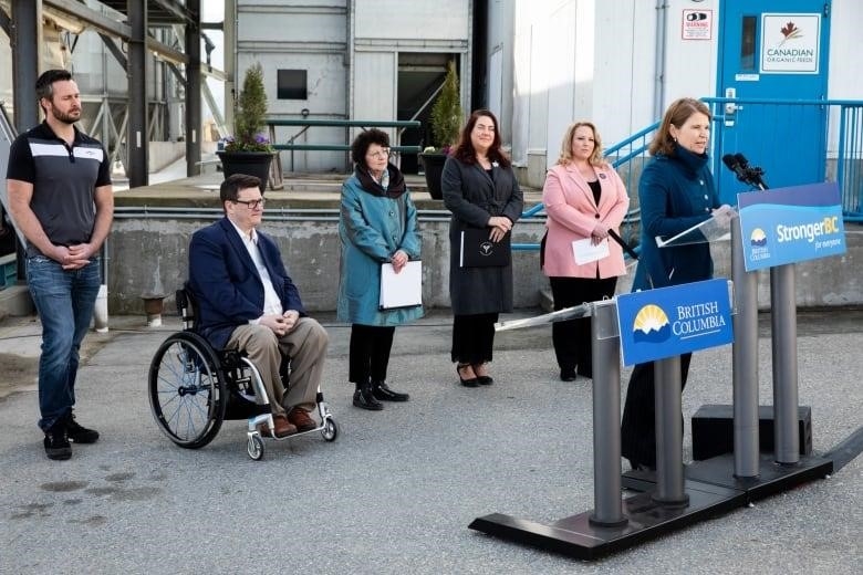 A woman wearing a blue jacket speaks at a podium marked 'Stronger BC', with two men and three women looking on behind her.