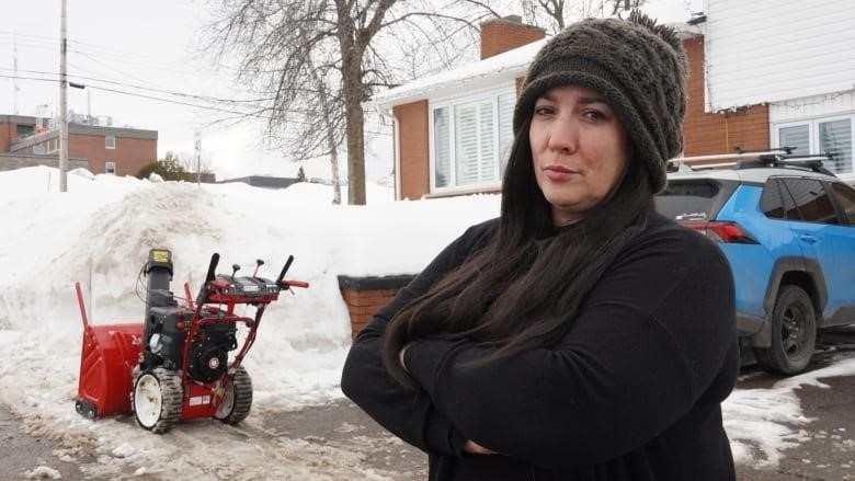 A woman crosses her arm in front of a snow plow and snow bank at a home.