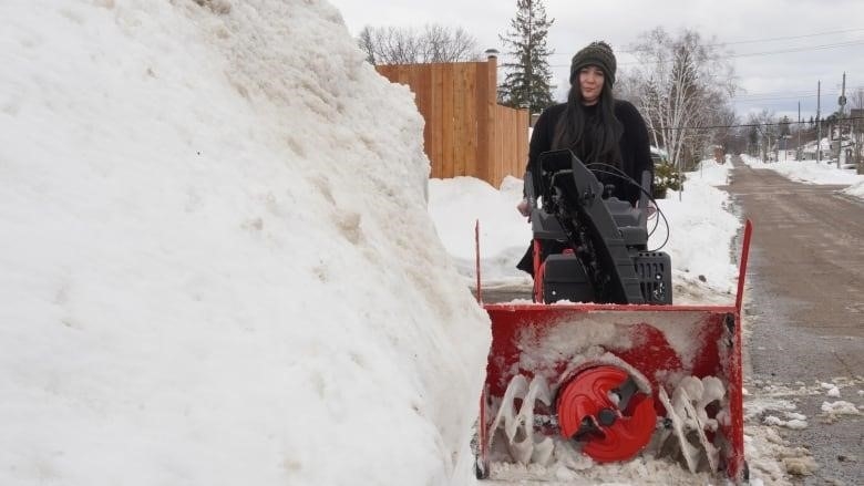 A woman stands next to a snowbank behind a snow blower.