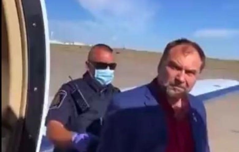 an officer wearing a medical mask handcuffs Artur Pawlowski on an airport tarmac. They are standing near an airplane.