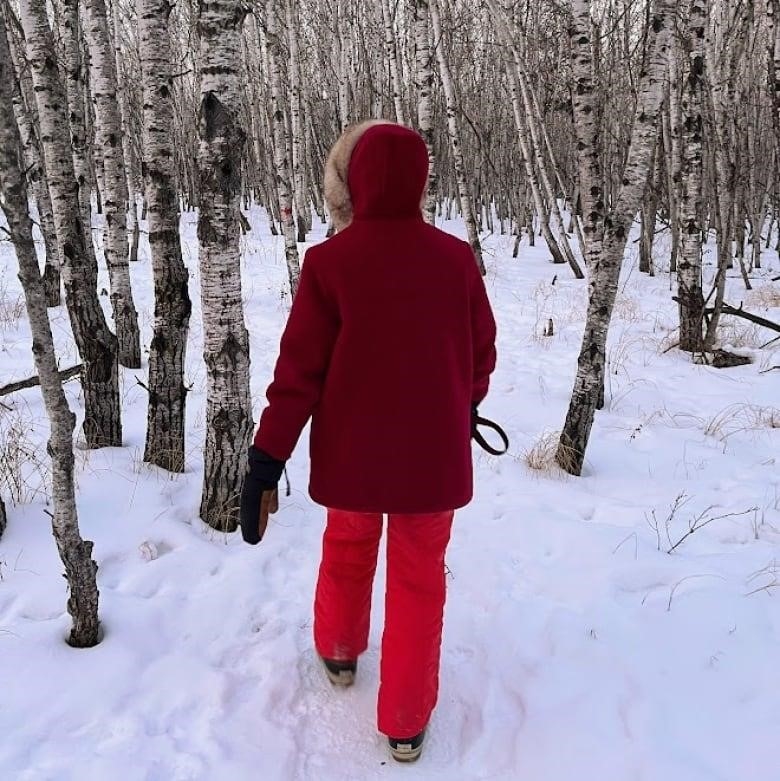 A person on a dark red winter coat and bright red snowpants walks on a snowy path through trees.