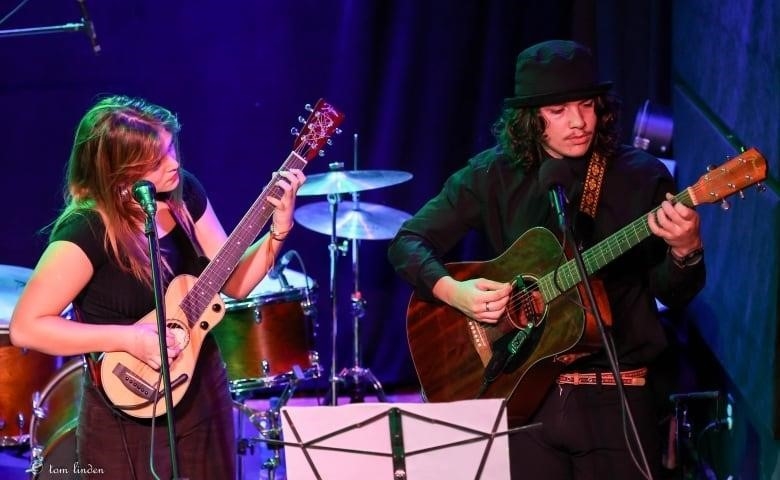 A man wearing all black plays a brown guitar on stage next to a young woman playing a smaller guitar. Behind the two is a drum set.