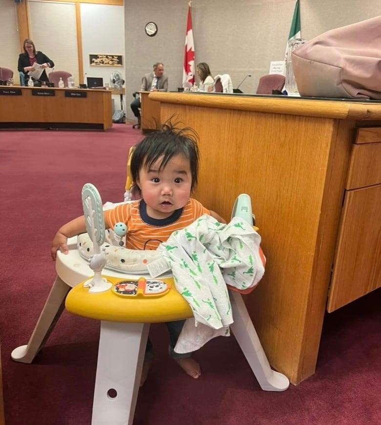 A baby sits in a play apparatus beside a desk inside a government meeting room.