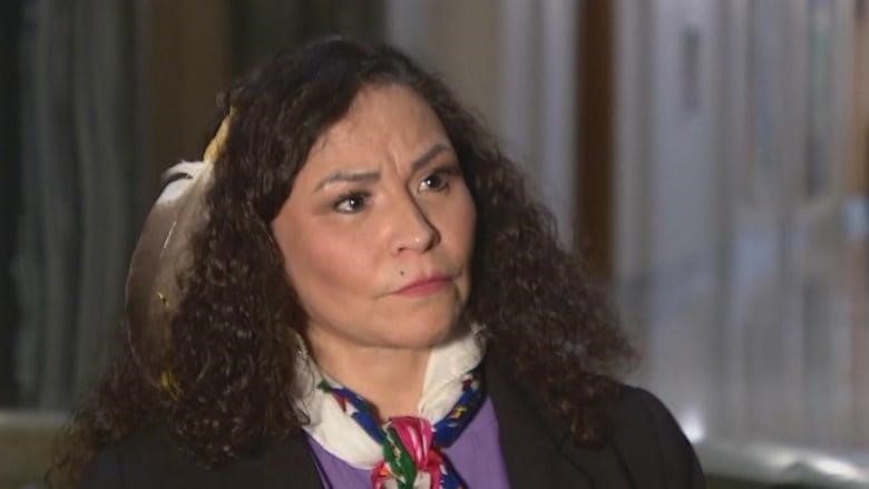 An Indigenous woman has a feather in her curly dark hair. She's wearing a colourful scarf around her neck, over a white dress shirt and black blazer.
