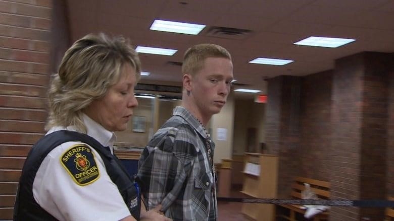 A red-haired man glaces to the camera. He is walking alongside a female sheriff's officer.