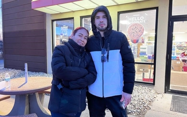 A woman leans her head on a younger man. Both standing outside a gas station.