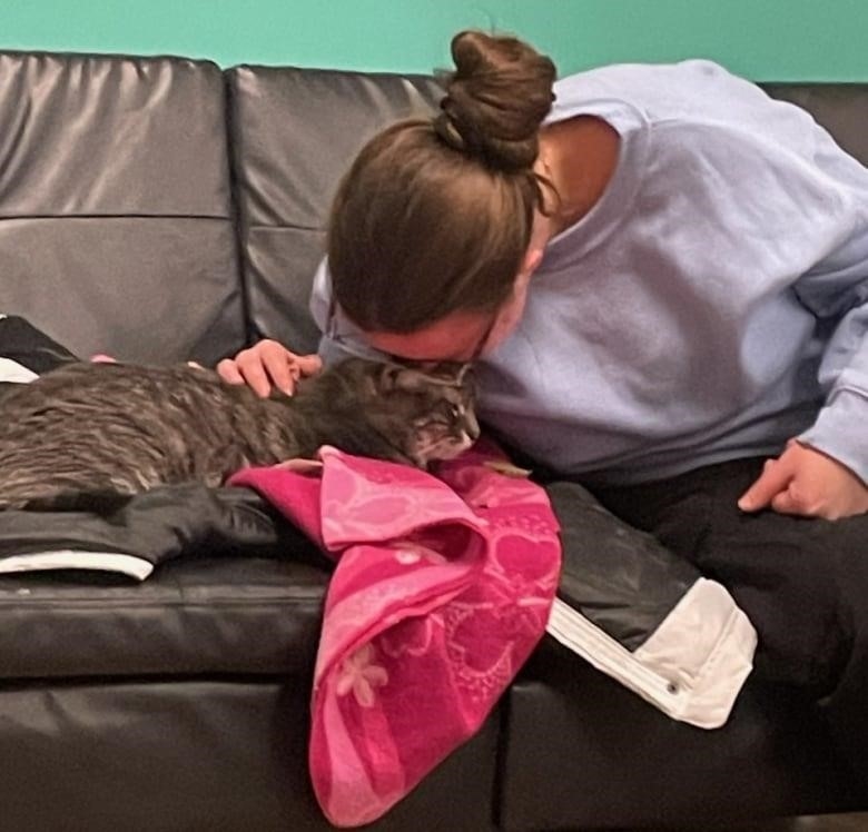 A woman with hair in a bun on top of her head and a grey lilac sweater kisses a grey tabby cat on a couch. The cat is sitting on a coat and a pink towel with flowers on it. 