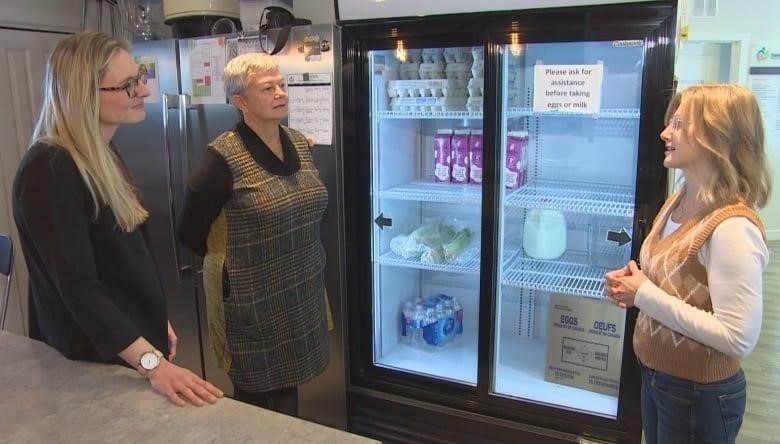 Three woman have a conversation in front of a commercial fridge.