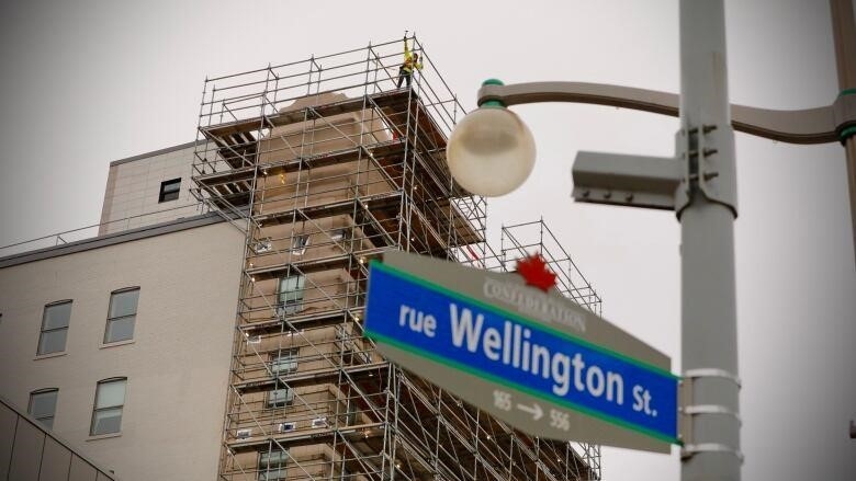A mid-rise building covered with scaffolding with construction workers on the scaffolding and a street sign 'rue Wellington St' in the foreground.