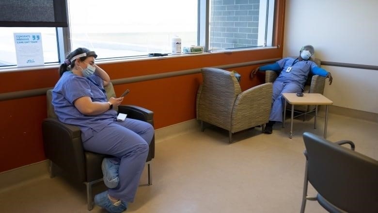 Two nurses collapse into chairs in a hospital lounge wearing scrubs.
