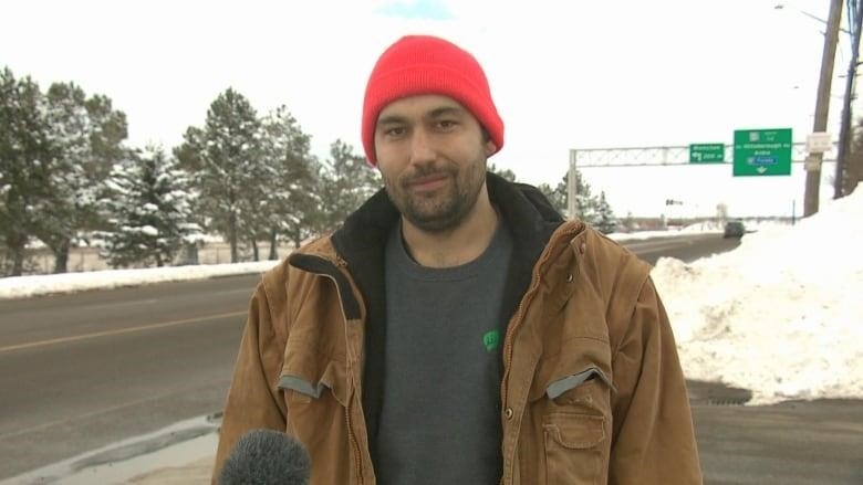 A bearded man with a red toque wearing an open brown coat stands with his back to a street. 
