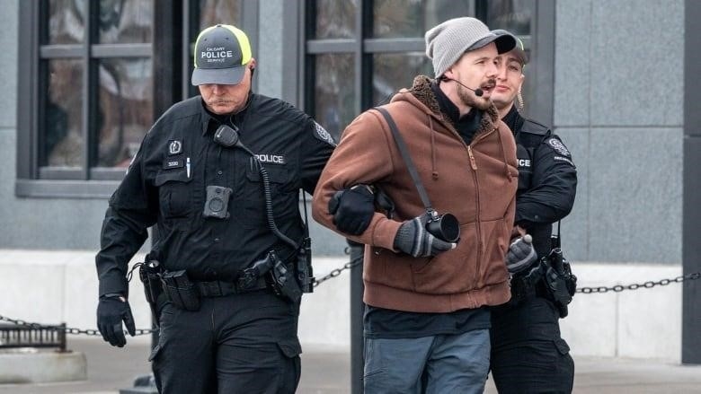 A man is flanked by two Calgary police officers, one on each arm, who appear to be marching him away from a building. 