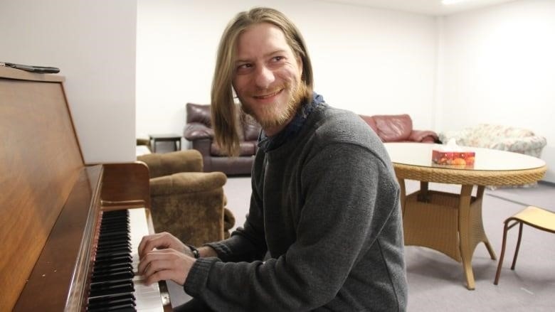 A man, Devin Edmiston, smiles while looking to his left as he sits on the piano and plays a song.