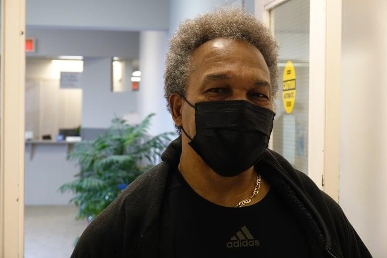 A Black man with curly grey hair stands in front of an open white door. He's wearing a black medical mask and a gold chain.