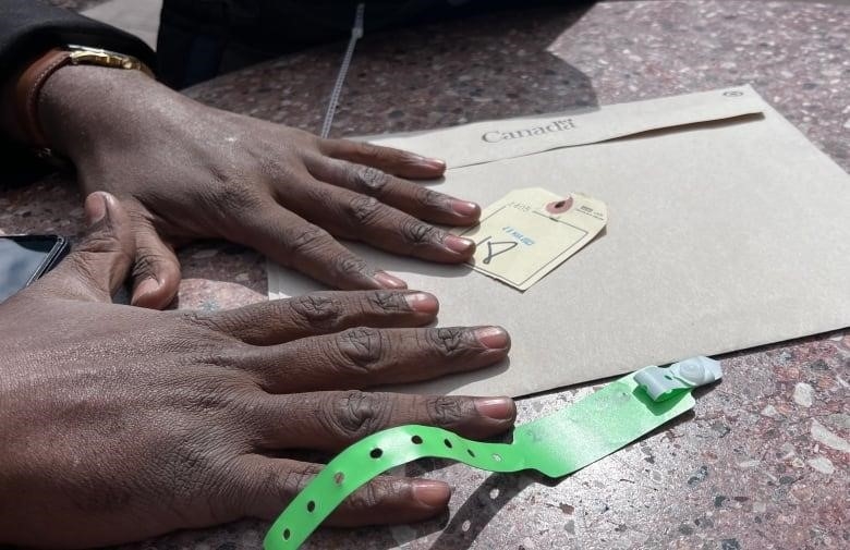 A man's hands over a brown Canadian government envelope. A tag with the number 18 on it and a plastic bracelet with numbers. 