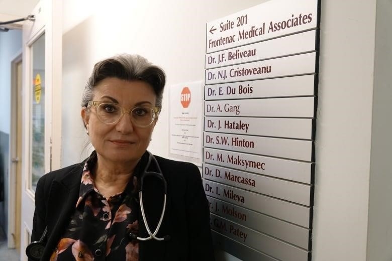 A woman with grey and black hair in a neat bun wears clear glasses and a stethoscope around her neck. She's standing next to a list of name cards with the titles of different doctors written in burgundy.