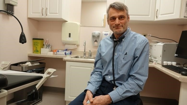 A man with grey hair wearing a blue and white-striped shirt sits in a small room surrounded by medical supplies. He's holding a blue surgical mask in his hand and has a stethoscope around his neck.