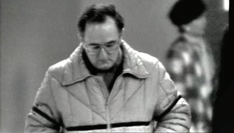 A black and white picture of a man walking with his head down. He has thinning dark hair and is wearing round glasses.