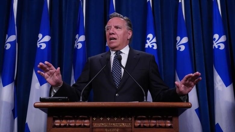 A man speaking at a podium in front of Quebec flags. 