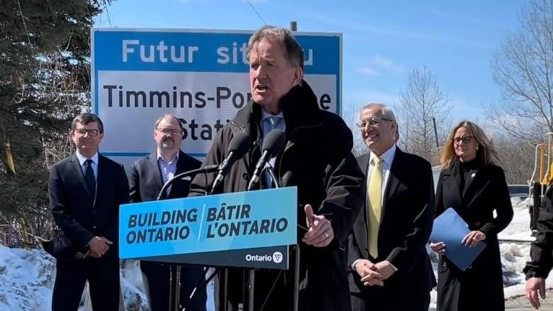 A politician stands at microphone and speaks, with others gathered behind him.