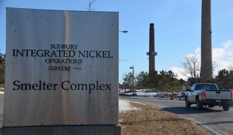 A pickup truck drives past a sign that reads 'Sudbury integrated nickel operations Glencore smelter complex' with two smoke stacks in the background. 