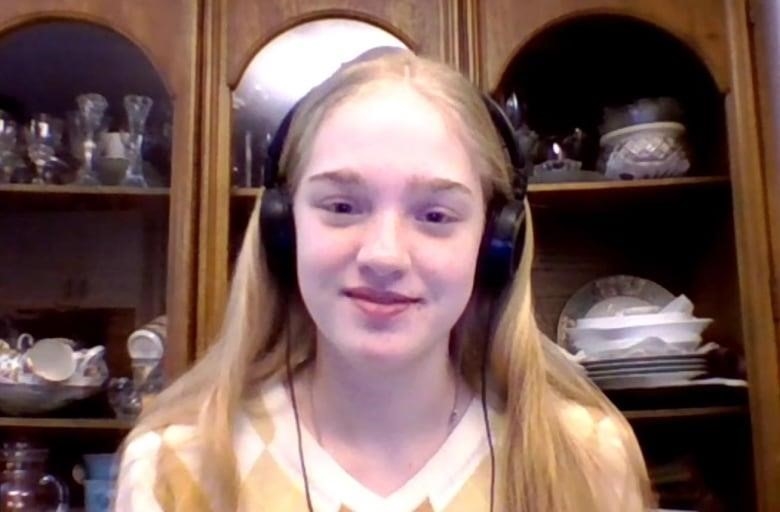 A teen girl with long blonde hair and wearing headphones shares a small smile in this screen capture from a video interview.  A dining room hutch is seen behind her. 