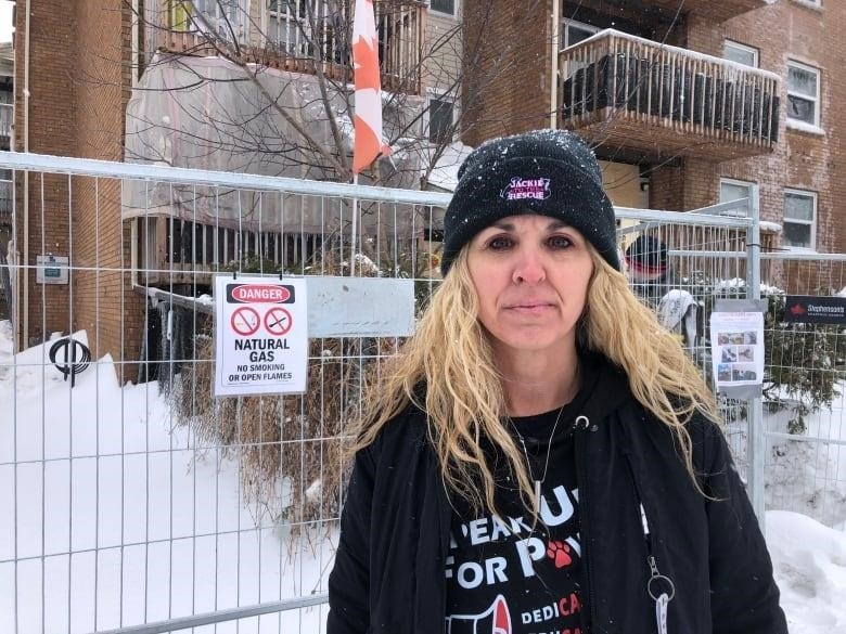 A woman with long, wavy blonde hair stands in front of fencing. She is wearing a black toque and black jacket over a black sweater. 