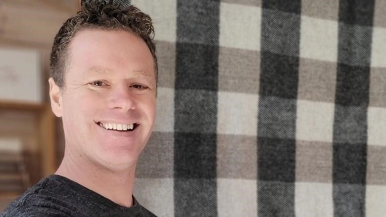 A man in a grey T-shirt smiles at the camera, while standing next to a wooly-looking blanket with green and grey squares on it.