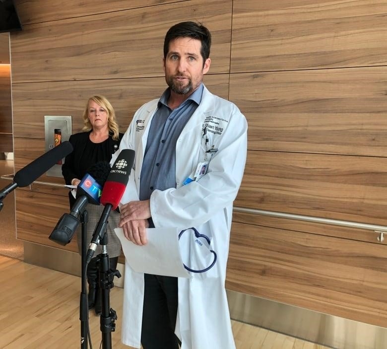 A woman with long blonde hair in a black top stands in the background as a man in a blue shirt and a lab coat with dark hair and a goatee stands behind three microphones.